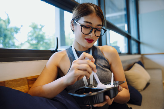 戴眼镜的亚洲女商人在办公室享用午餐盒。