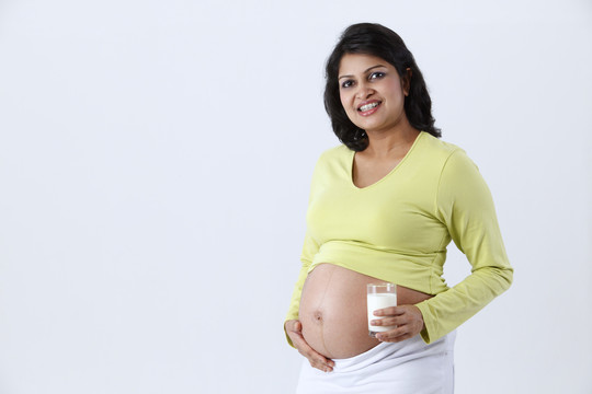 微笑着的孕妇站着，拿着一个杯子，杯子里的牛奶被隔离在灰色的背景上。