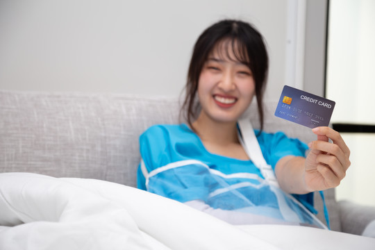 在医院里，一名持有保险信用卡的亚洲妇女坐在床上，手臂上穿着病人服，戴着柔软的夹板