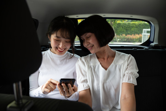 亚洲年轻女性和老年女性在汽车后座使用智能手机，乘客使用应用程序预订乘车和点对点乘车共享概念