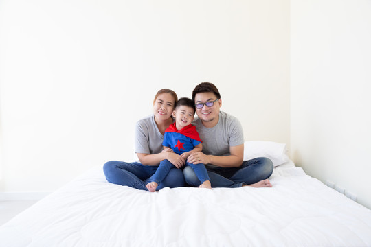 笑容满面的亚洲家庭和穿着超级英雄套装的儿子坐在卧室的白色床上