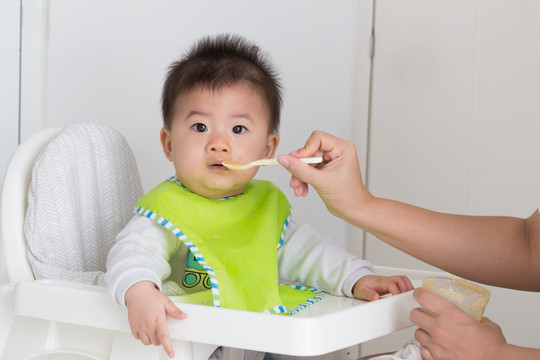 快乐的孩子坐在椅子上，妈妈在家里用勺子喂男孩，亚洲模特