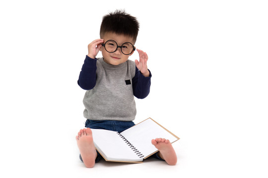 一个可爱的亚洲小男孩戴着眼镜坐在地板上读一本白色背景下的书，回到学校的概念，1岁10个月大