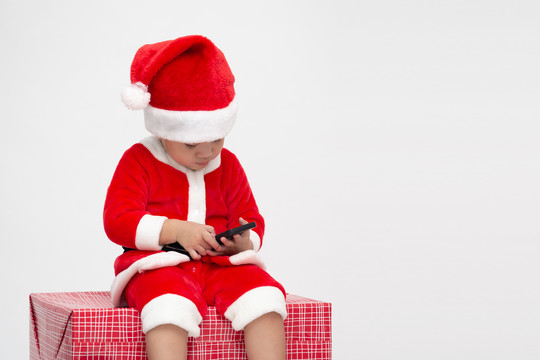 穿着圣诞老人套装的亚洲男婴，手持手机，坐在白色背景的礼品盒上