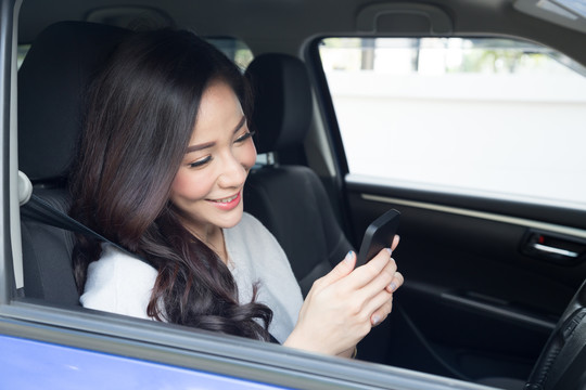 年轻美丽的亚洲女性坐在车里的驾驶座上，使用带有移动应用程序或地图gps导航、触摸屏的智能手机，阅读聊