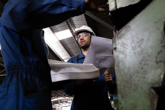 工厂工程师阅读机器操作手册，穿着蓝色工作服和安全帽站立。重工业工厂机械技术专家概念