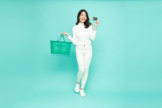 浅绿背景上，一位手持购物篮、展示信用卡、面带微笑的快乐亚洲女性