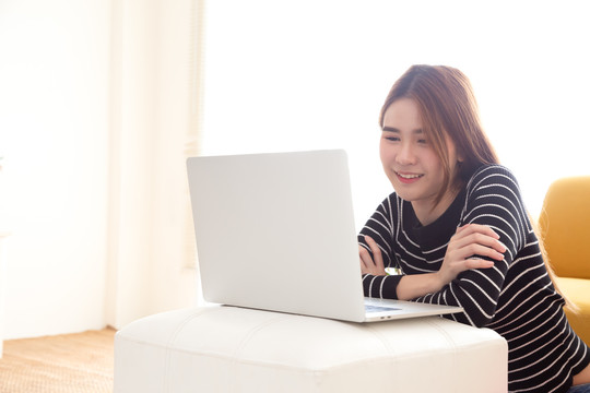 年轻的亚洲女性坐在地板上，在家里客厅里用笔记本电脑在线观看电影、电视