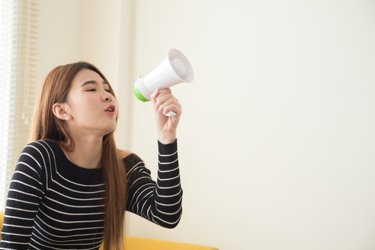 美丽的亚洲年轻女子在家里客厅里用扩音器发布公告或交流，来自穿着黑色衬衫坐在黄色沙发上的少年的信息发布