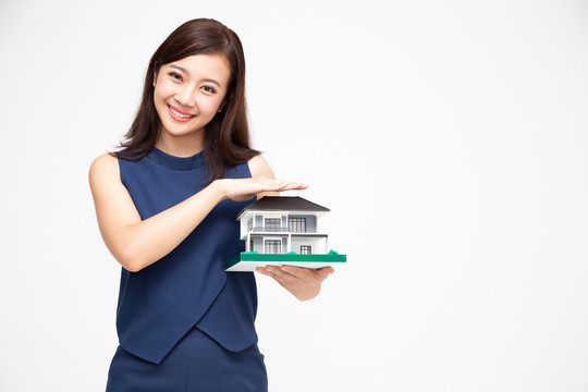 美丽的年轻亚洲女性肖像，双手保护房屋或家庭模型，白色背景，房地产和家庭保险概念