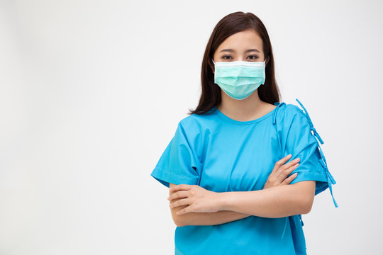 年轻的亚洲女性，穿着患者服装，双臂交叉，戴着白色背景隔离的面罩