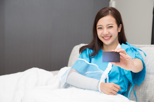 在医院的病床上，一名亚洲妇女拿着保险卡，手臂上穿着病人服，戴着柔软的夹板