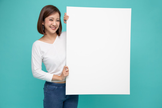 微笑快乐的亚洲妇女，站在浅绿背景上孤立的白色大海报后面