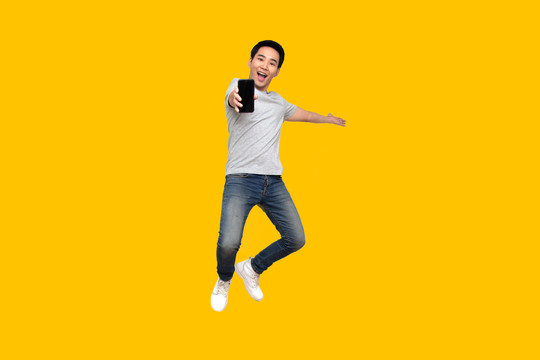 亚洲男子在黄色背景上跳起并展示手机