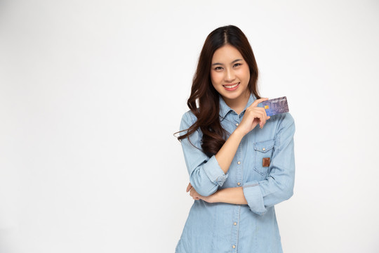 照片中的亚洲女性穿着蓝色牛仔裤衬衫，手持信用卡，微笑隔离在白色背景上，年轻女性微笑着看着相机，快乐的