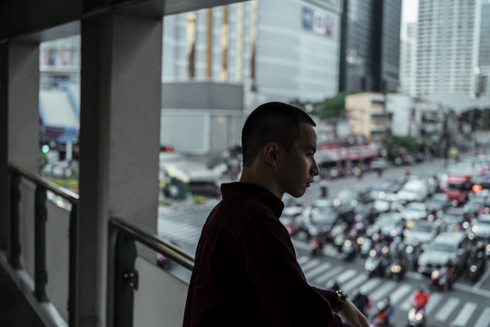 身穿红衫的光头男子站在满是汽车和摩托车的道路上方的天桥上。