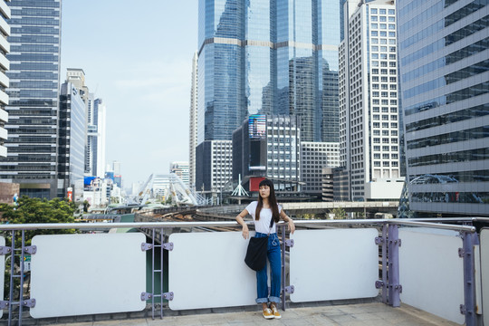 一个留着刘海发型的可爱泰国女人靠在扶手上，等着有人来接她。城市景观背景。