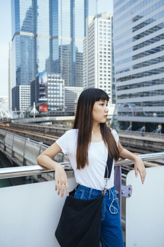 一个留着刘海发型的可爱泰国女人靠在扶手上，等着有人来接她。城市景观背景。