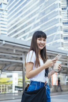留着刘海发型的年轻亚洲女孩手持智能手机和一杯泡泡茶，通过耳机听音乐。