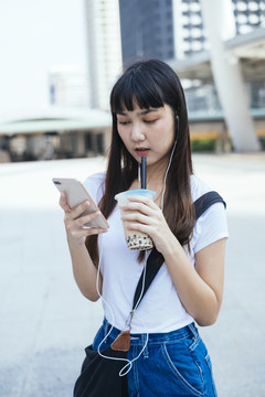 一位留着刘海发型的可爱亚洲年轻女子一边喝泡泡茶一边玩智能手机。