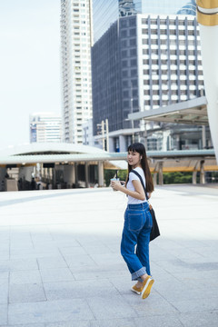 穿着白色衬衫和蓝色牛仔裤的年轻亚洲女性手拿泡泡茶在城里散步。