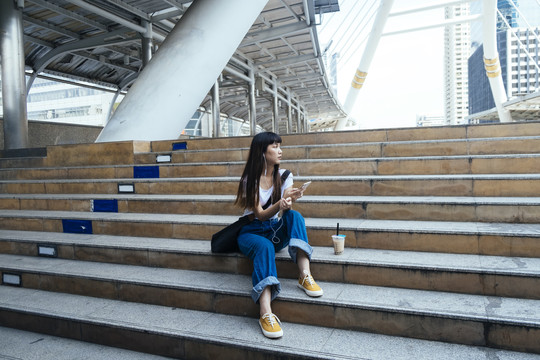 亚洲年轻的泰国女子坐在楼梯上，玩着智能手机，用耳机听音乐，等待着她的男朋友。