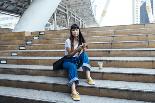亚洲年轻的泰国女子坐在楼梯上，玩着智能手机，用耳机听音乐，等待着她的男朋友。