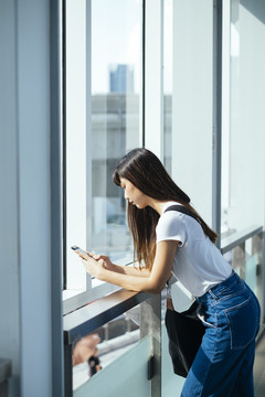泰国年轻女士在大楼镜子前使用智能手机的侧视图。聊天和上网。