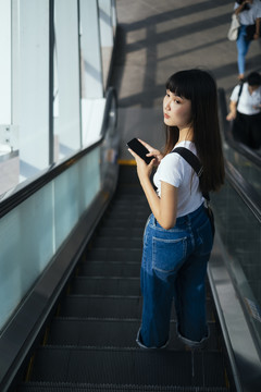 一位留着刘海发型的美丽亚洲年轻女子手持智能手机站在自动扶梯上。