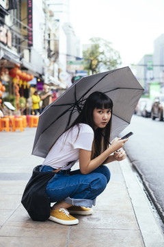 可爱的泰国年轻女子手持雨伞和智能手机坐在街上。