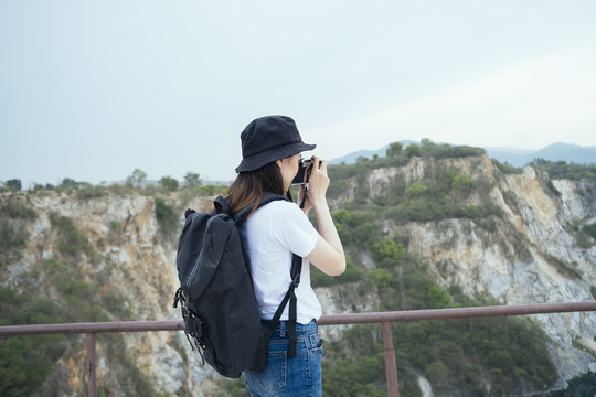 后视图-年轻的亚泰黑头发旅行家妇女在山区用相机拍照。