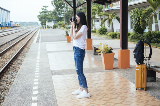 侧视图-年轻的亚泰黑头发旅行家妇女在火车站用相机拍照。