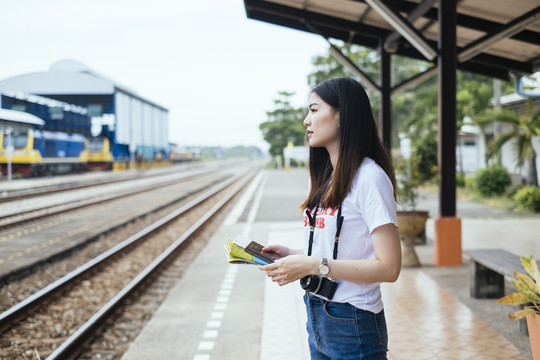 在火车站月台上拿着纸质地图的年轻亚裔泰国长黑发女旅行者。