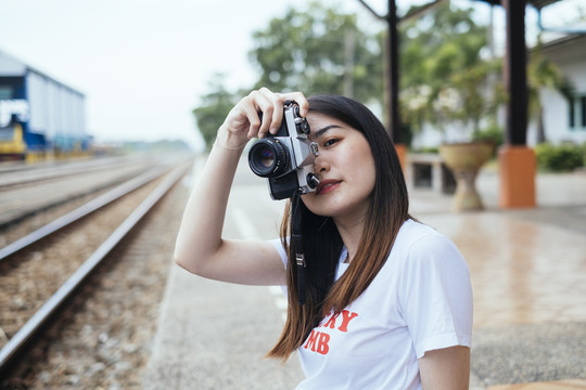 年轻的亚裔泰裔留着深色长发的女旅行家摄影师手持相机坐在火车站月台上。