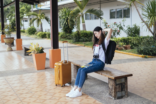 一位年轻的亚裔泰国长毛黑发女旅行者坐在长凳上等火车。