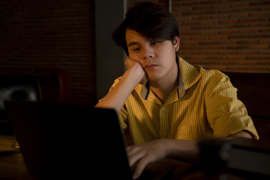 无聊的亚洲年轻人晚上在办公室里用数字笔记本电脑工作。