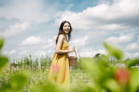 侧视图-身着黄色连衣裙的亚洲美女手持篮子在田野中行走。