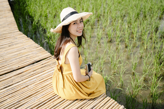 穿着黄色衣服的亚洲旅行家美女手持相机坐在稻田的木桥上。