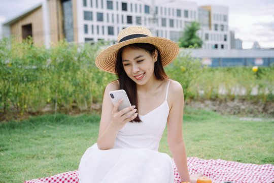 美丽的亚洲旅行家，穿着白色连衣裙，戴着白色帽子，在户外公园使用智能手机。
