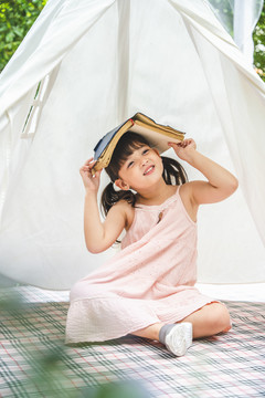 可爱的亚洲小女孩，头上戴着一本书，呆在花园里的白色露营帐篷里。