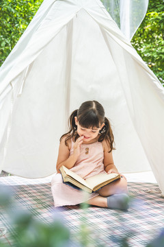 可爱的亚洲小女孩在户外花园的白色露营帐篷里看书。