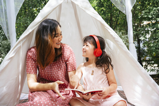 母亲和女儿呆在白色的露营帐篷里，在户外花园里读着一本书中的故事。