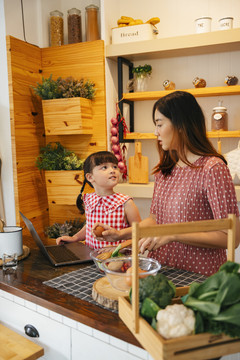 亚洲小孩和妈妈在厨房里互相帮助做饭。人们在网上搜索食物配方。
