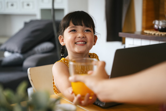 亚洲小女孩一边用笔记本电脑一边从妈妈那里拿一杯橙汁。