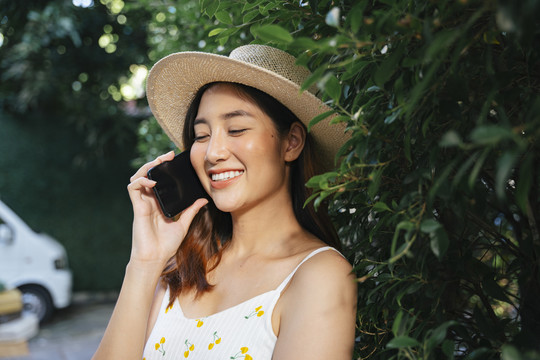 美丽的亚洲女游客戴着帽子，穿着白色衣服在公园里打电话。