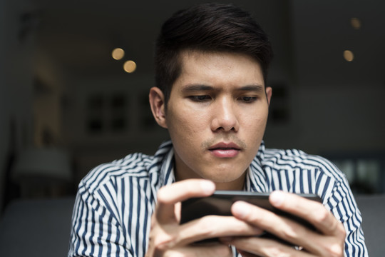 一名亚洲男子坐在沙发上，在家里用智能手机玩手机游戏。