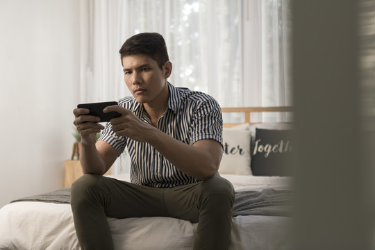 亚洲男人在家里的卧室里用智能手机玩手机游戏。
