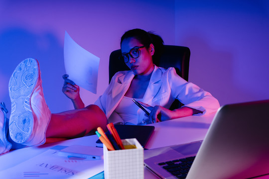 时髦的亚洲女商人把腿放在办公桌上。霓虹色灯光色调。