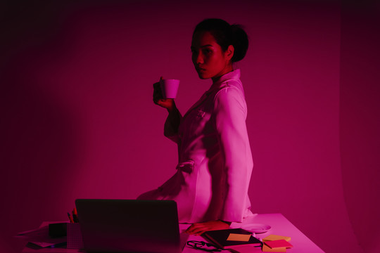 在红色霓虹灯下，女商人手持一杯咖啡坐在办公桌上的肖像。