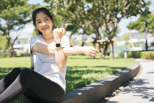 运动型亚洲女性在公园慢跑前伸展手臂热身。